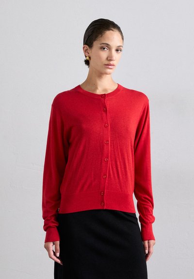 Rød langærmet cardigan lavet af blødt strikmateriale med rund halsudskæring og knaplukning. Stylet over en sort nederdel.
