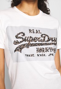 Bílý bavlněný tričko s grafickým motivem stříbrné barvy a textem "REAL SuperDry" v leopardím vzoru. Krátké rukávy a volný střih.
