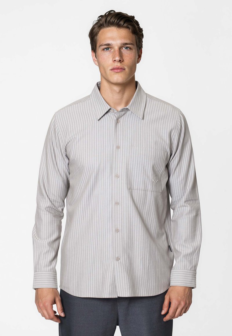Jeune homme aux cheveux bruns courts portant une chemise à manches longues rayée gris clair boutonnée et un pantalon foncé, debout devant un fond blanc uni.