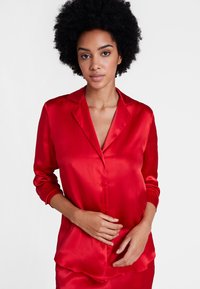 Femme aux cheveux bouclés naturels portant une chemise rouge en satin à boutons et un bas assorti sur fond blanc uni.