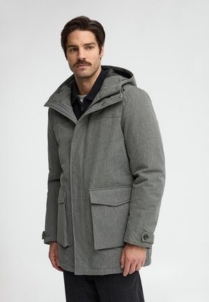 Hombre con cabello castaño y bigote que lleva un abrigo de invierno gris con capucha y grandes bolsillos delanteros sobre un fondo claro liso.