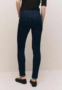 Jeans skinny azul oscuro con una alta cintura, bolsillos traseros y una textura suave. Llevados con zapatos planos negros, con un dobladillo enrollado.