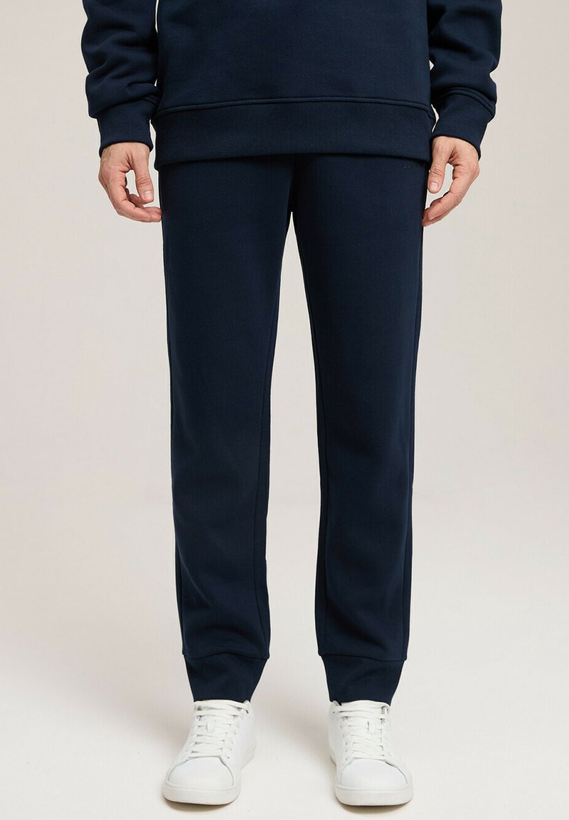 Granatowe spodnie dresowe z miękkiego materiału o tapered designie. Posiadają elastyczny pas i mankiety, zestawione z białymi trampkami.