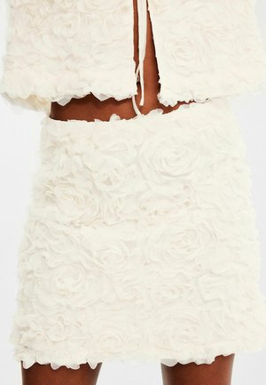 Witte twee-delige outfit met een cropped jacket en een aansluitende rok, beide versierd met textuurpatronen van bloemen gemaakt van gelaagd stof.