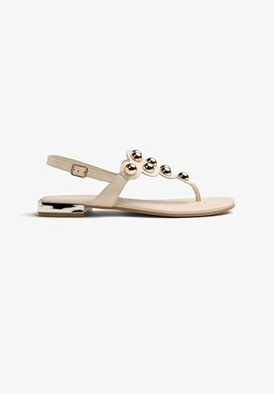 Sandalia plana beige con tira trasera, adornos metálicos dorados en forma de bola en la tira frontal y tacón cuadrado bajo con detalle dorado.