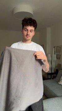 Tessuto grigio, leggermente spiegazzato, tenuto da una persona. Il capo sembra morbido con un drappeggio rilassato, presenta un collo rotondo e nessun motivo visibile.