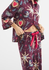Paarse pyjama-set met een colbertachtige shirt en broek met elastische tailleband, versierd met kleurrijke prints van harten, vogels en zonmotieven. Rode accenten.