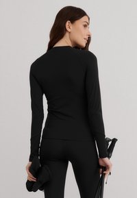 Camiseta deportiva negra de manga larga con un diseño ajustado, textura suave y mangas raglán, combinada con leggings negros y sosteniendo bastones de senderismo.