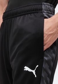 Άτομο που φοράει μαύρη φόρμα Puma με το χέρι στην τσέπη· το παντελόνι έχει ζιγκ-ζαγκ πλαϊνή ρίγα και εμφανές λευκό λογότυπο Puma.