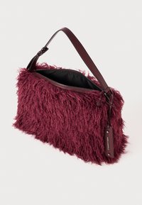 Burgunderfarbene Tasche mit flauschiger Textur, gestaltet mit einem Reißverschluss und einem braunen Ledertrageriemen. Auffälliges Etikett auf der Seite.