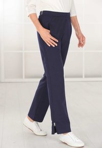 Marineblauwe broek met elastische tailleband, zijzakken en ritsen bij de enkels, gemaakt van zachte stof, gecombineerd met witte sneakers op een lichte vloer.