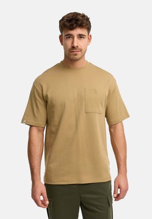 Młody mężczyzna w beżowej koszuli z krótkim rękawem i kieszenią na piersi oraz ciemnozielonych spodniach bojówkach, stojący na tle gładkiej powierzchni.