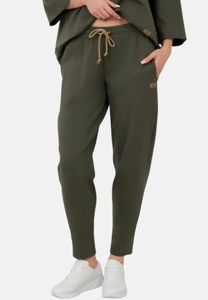 Osoba nosząca oliwkowe spodnie dresowe z troczkiem w talii, zestawione z białymi trampkami; jedna ręka w kieszeni, druga poprawiająca dół koszuli.