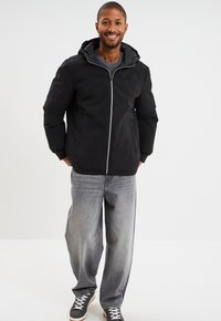 Chaqueta negra con capucha, cierre frontal, dos bolsillos laterales y forro gris. Combinada con pantalones anchos de mezclilla gris claro y zapatillas negras.
