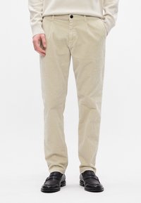 Calças de veludo cotelê bege com corte reto, apresentando bolsos frontais, um fecho de botão único e uma textura subtil. Combinadas com sapatilhas pretas.