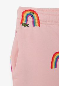 Pantalones de chándal rosas con arcoíris bordados y un logo de cocodrilo verde. La tela parece suave y con una textura lisa.