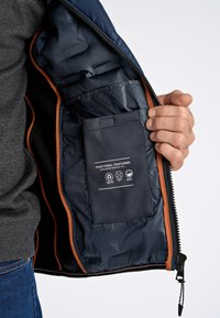 Marinblå pufferjackas insida med orange detaljer, som visar en strukturerad ficka märkt "FUNKTIONSFUNKTIONER" med vårdsymboler. Dragkedjestängning.