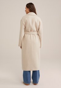 Beige ullblandad kappa med bred krage, långa ärmar och midjebälte. Har en rak silhuett och går nedanför knäna.