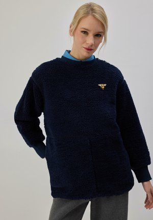 Femme blonde portant un pull texturé bleu marine avec un petit emblème d'abeille sur la poitrine et un pantalon gris sur un fond uni.
