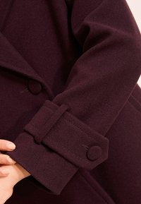 Manica di un cappotto di lana viola scuro con polsino cucito e bottone rivestito in tessuto rotondo, tenuta da una mano con unghie naturali.