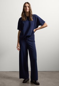 Jonge vrouw staat met een hand op de heup, gekleed in een lose marineblauwe top en wijde broek, gecombineerd met zwarte loafers, tegen een effen grijze muur.