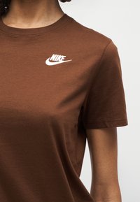 Άτομο με καφέ μπλουζάκι Nike με λευκό λογότυπο Nike στο αριστερό στήθος, εμφανίζεται από τον λαιμό έως το μέσο του κορμού σε απλό φόντο.