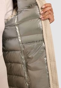 Beežid täidisega jope sisevoodriga metallik-hõbedases toonis, millel on tikitud tekstuur ja küljetasku. Sile kangas kontrastib välimusega.