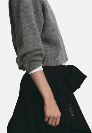 Persona vistiendo un suéter de punto gris con mangas remangadas sobre una camiseta blanca, combinado con una falda negra con un lazo y botones.
