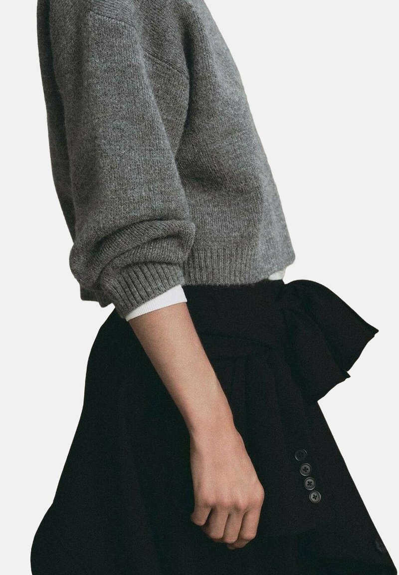 Persona che indossa un maglione lavorato a maglia grigio con le maniche arrotolate sopra una camicia bianca, abbinato a una gonna nera con un fiocco e bottoni.