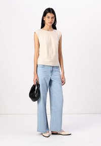 Beige ærmeløs top med høj hals, parret med lyseblå brede bukser. Accessories inkluderer en sort taske og beige tofarvede sko.