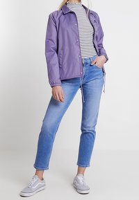 Světle fialová zipová bunda s patentkami, kombinována s bílým a černým pruhovaným rolákem, modrými džíny a šedými teniskami.