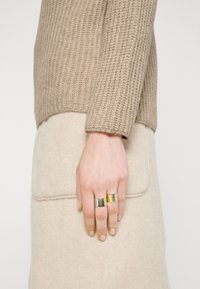 Pull beige en tricot avec un motif texturé, associé à un tissu beige doux en dessous. Trois anneaux métalliques en argent, or et une teinte plus foncée.