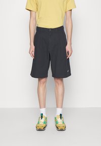Person wearing black knee-length shorts, yellow T-shirt, white socks, and yellow and green sneakers, standing against a plain white background.