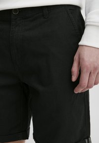 Fekete pamut rövidnadrág laza fittel, egyenes szabású, felgöndörített alsó szegéllyel és gombos záródással. A felülete simának tűnik, minimális varrással.