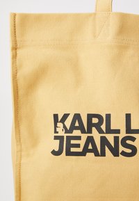 Geltona kanvos pirkinių maišelis su dideliu juodu "KARL L JEANS" logotipu ir grafiniu akcentu. Matoma tekstūruota medžiaga ir tvirtas siuvimas.