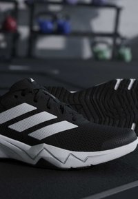 Κοντινή λήψη σε ασπρόμαυρα αθλητικά παπούτσια Adidas που ξεκουράζονται σε ένα σκοτεινό δάπεδο γυμναστηρίου με θολό εξοπλισμό γυμναστηρίου στο φόντο.
