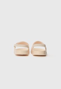 Sandali beige chiari con una superficie liscia, dotati di una soffice fascia e un design aperto all'altezza delle dita. La suola è piatta e minimalista nella forma.