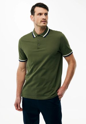 Mann trägt olivgrünes Poloshirt mit weißem und marineblauem gestreiftem Kragen und Ärmelbündchen, steht mit einer Hand in der Tasche und schaut nach rechts.