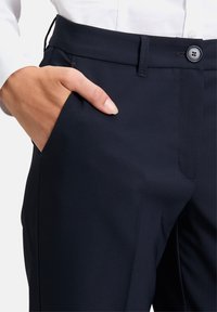 Marina anpassade byxor visas från midjan och ner; har en slät yta, sidofickor och en knappstängning vid midjan.