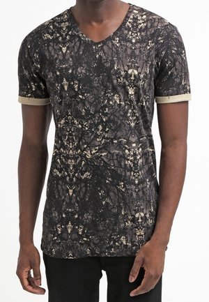 Mężczyzna noszący czarno-beżowy T-shirt z abstrakcyjnym wzorem i dekoltem w serek, z podwiniętymi beżowymi mankietami oraz czarnymi spodniami na białym tle.