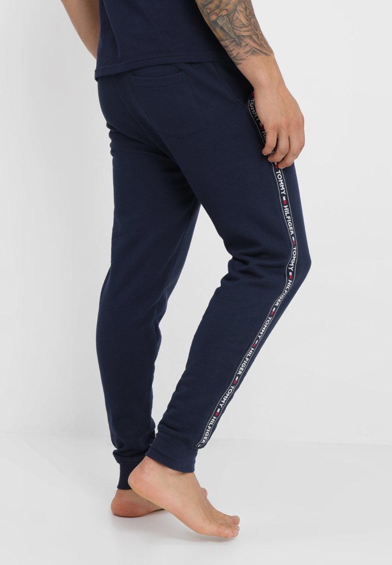 Osoba ubrana w granatowe dresy Tommy Hilfiger z paskiem z logo po boku, stojąca na jednej nodze boso, z widocznym tatuażem na ramieniu.