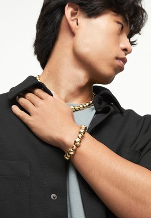 Collier et bracelet dorés composés de perles rondes, portés par une personne vêtue d'une chemise noire sur un fond neutre.