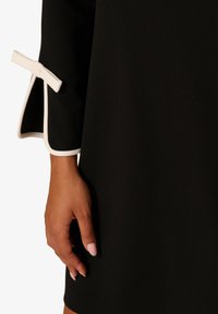 Černé šaty s holubími rukávy, zdobené bílým lemem a mašlí. Hladký materiál s přiléhavým střihem. Ruka umístěna na lemu.