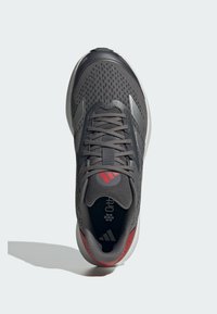 Zapatilla atlética gris con parte superior de malla, cierre de cordones y acentos rojos en el talón. Cuenta con un cuello acolchado y suela texturizada para mayor tracción.