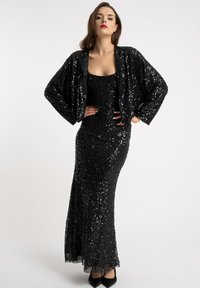 Czarna sukienka z cekinami o dopasowanym kroju, w zestawieniu z pasującą krótką kurtką. Obie mają błyszczącą teksturę i pełnej długości rękawy.