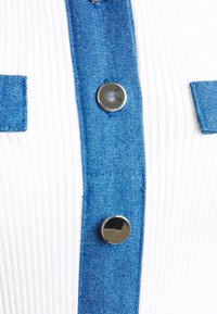 Tissu côtelé blanc avec des accents en denim bleu, présentant un devant boutonné et des détails de poche. Comprend des boutons en métal argenté pour la fermeture.