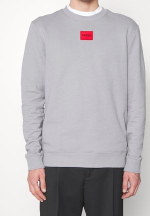 Grauer Pullover aus einem Baumwollmischgewebe, mit langen Ärmeln, gerippten Bündchen und einem roten rechteckigen Logopatch auf der Brust.