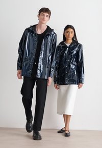 Marineblauwe, glanzende regenjacks met capuchons, voorzien van drukknoopsluitingen en zakken, gedragen over zwart-witte casual outfits.