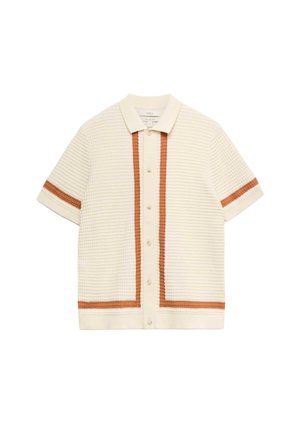 Hombre con cárdigan corto de manga corta color crema con textura y rayas marrones sobre camiseta blanca y pantalones beige con cordón, mirando hacia abajo y hacia un lado.