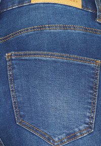 Jeans de ganga azuis com um bolso traseiro com costura amarela, textura suave e uma etiqueta de marca em couro na cintura.